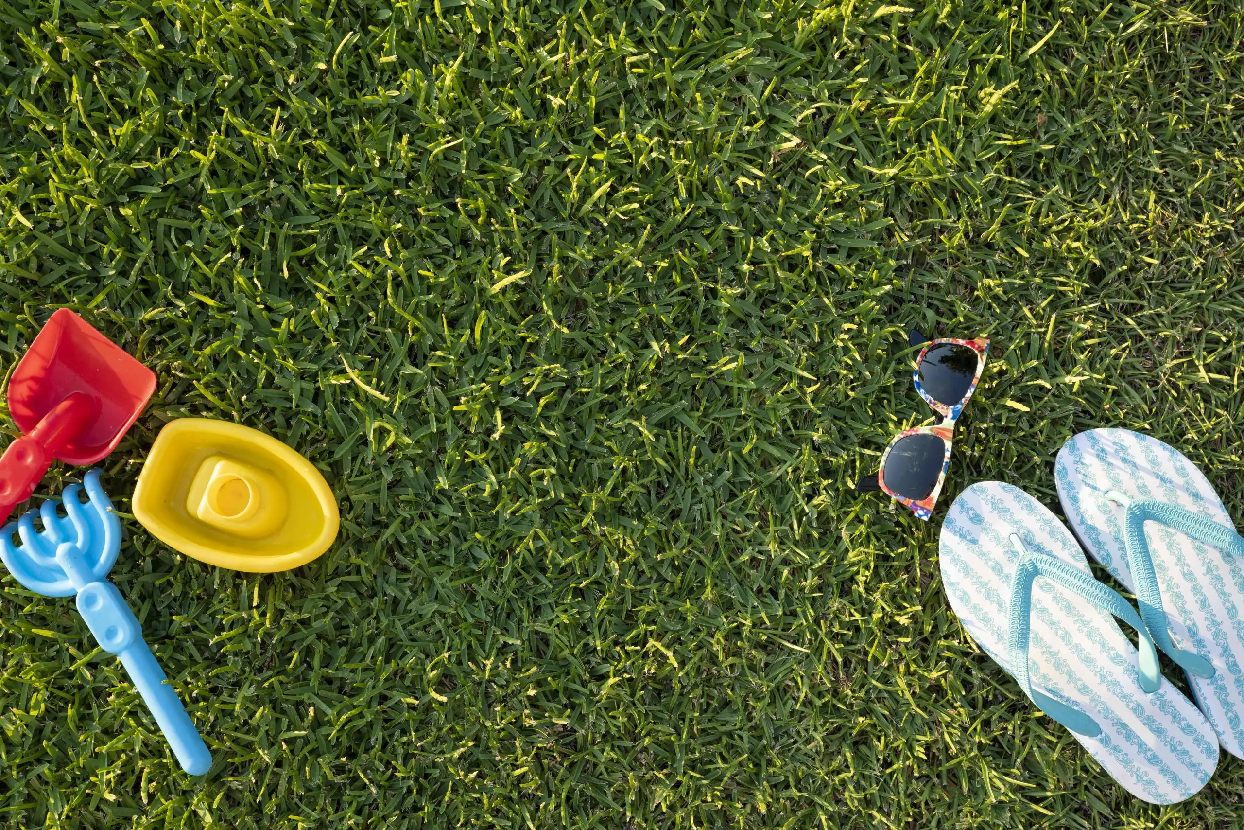 Overhead view of a thick, evenly cut green lawn with sandals, sunglasses and children's sand toys resting on the grass
